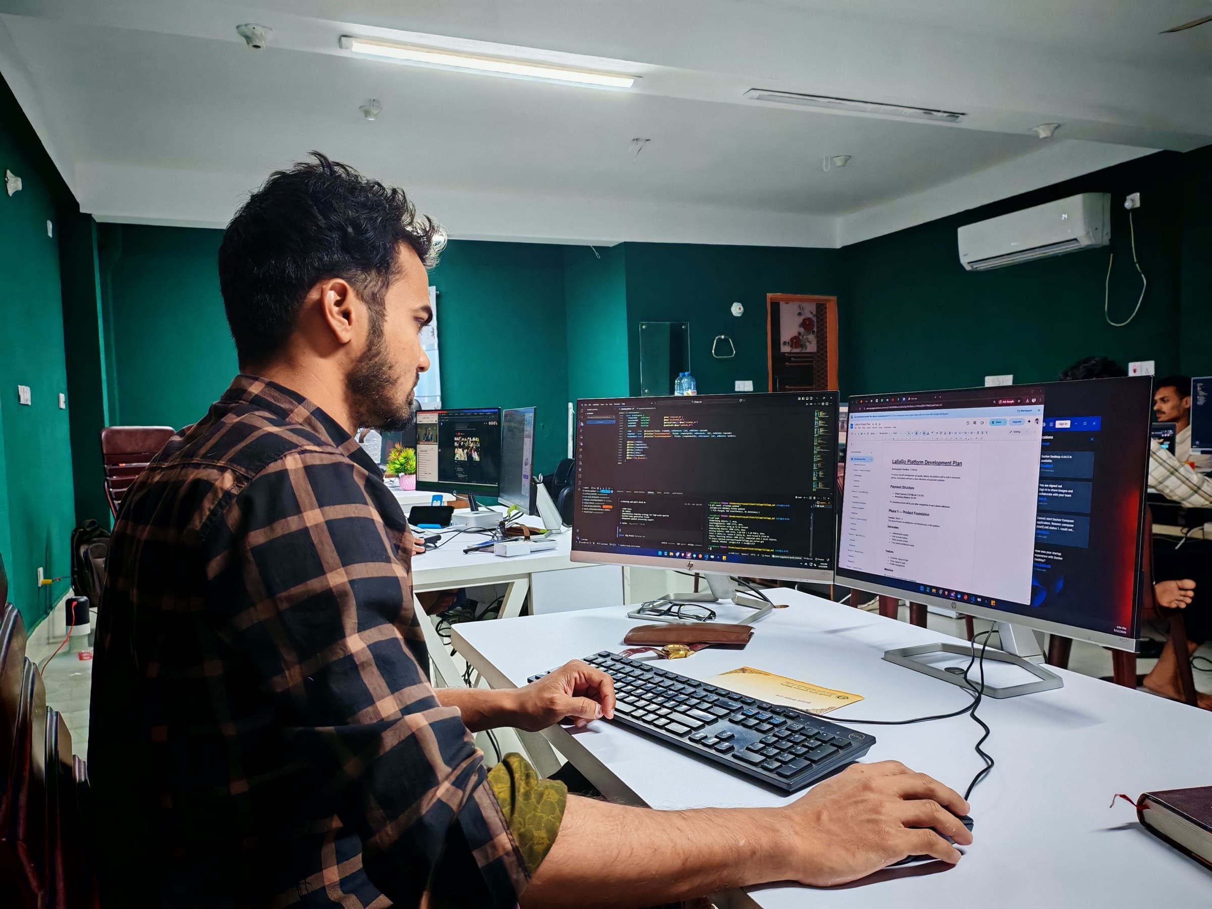 A developer at a dual-monitor desk with a code editor and a project plan open — full stack development work in 2026