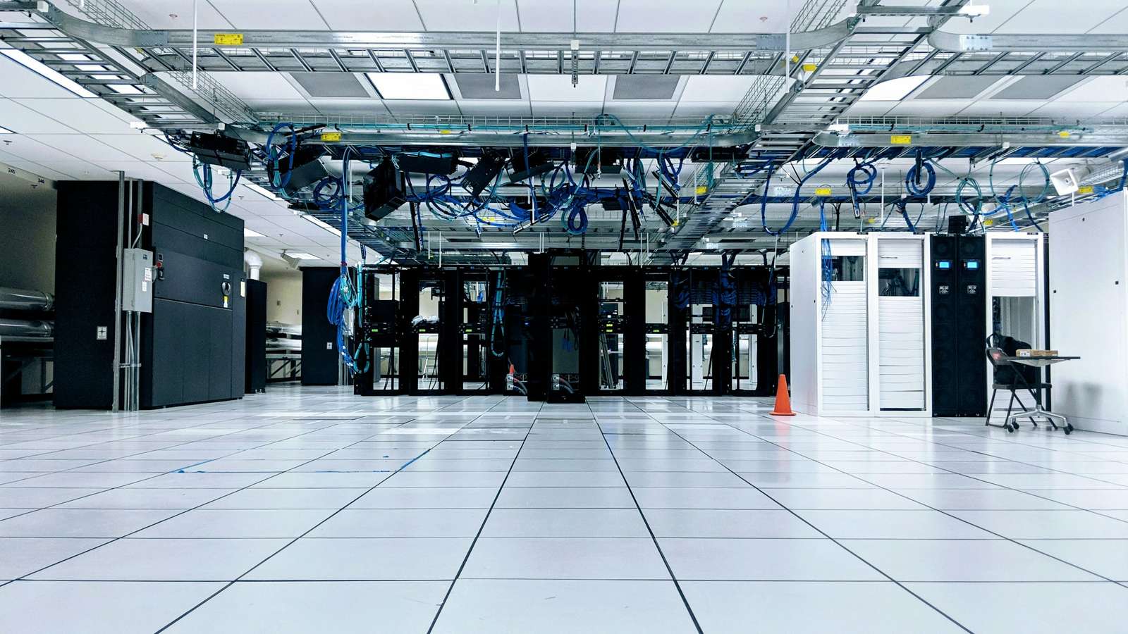 A data centre floor with rows of black server cabinets under overhead trays of blue network cable, on a white raised tile floor. CDN points of presence are rooms like this one, placed in cities around the world