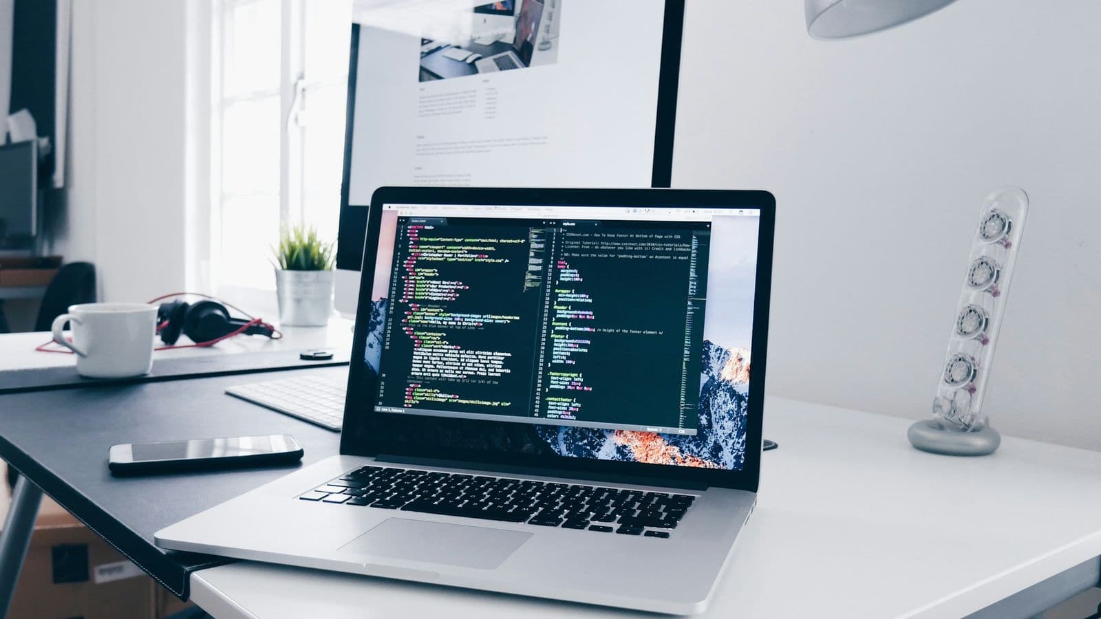 A MacBook on a white desk showing lines of code in a dark editor, next to a coffee cup, a smartphone, a small potted plant and a larger monitor behind. Good cloud architecture is decided at a desk like this, long before the first customer signs in