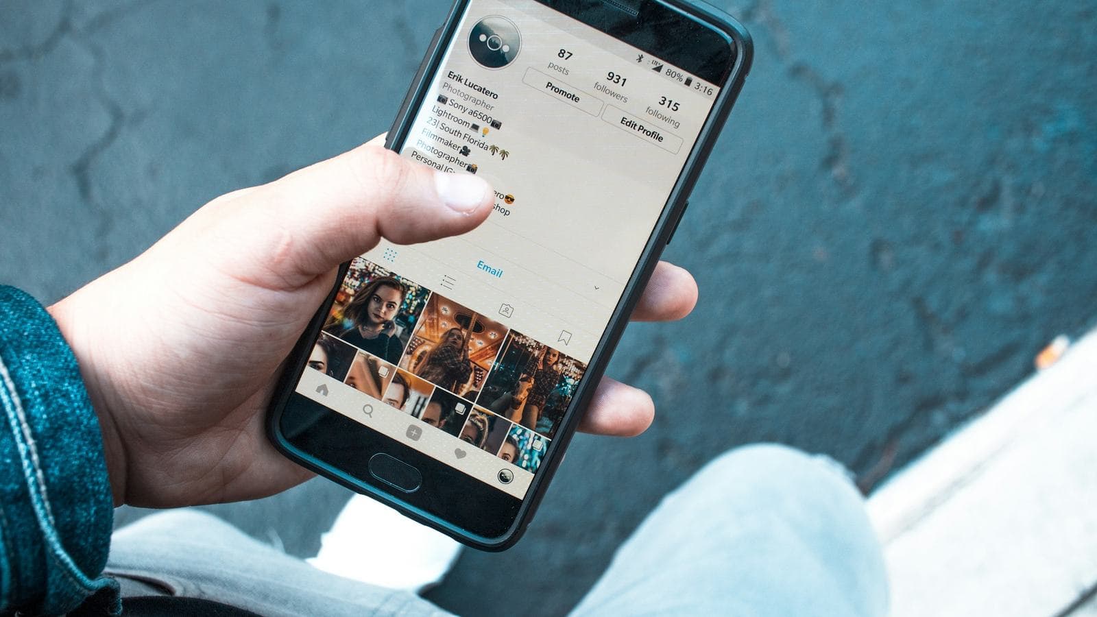 A hand holding an Android phone showing an Instagram profile grid of photos, with denim jeans and a pavement in the background. Posting a photo is one of the most ordinary cloud computing tasks there is