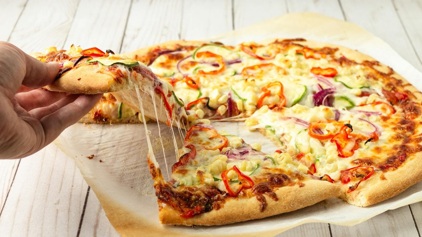 A hand lifting a slice of vegetable pizza with stretching cheese, topped with peppers, onions and courgette, on parchment paper on a white wooden table. How much of this pizza you make yourself is a surprisingly good way to understand the cloud service models