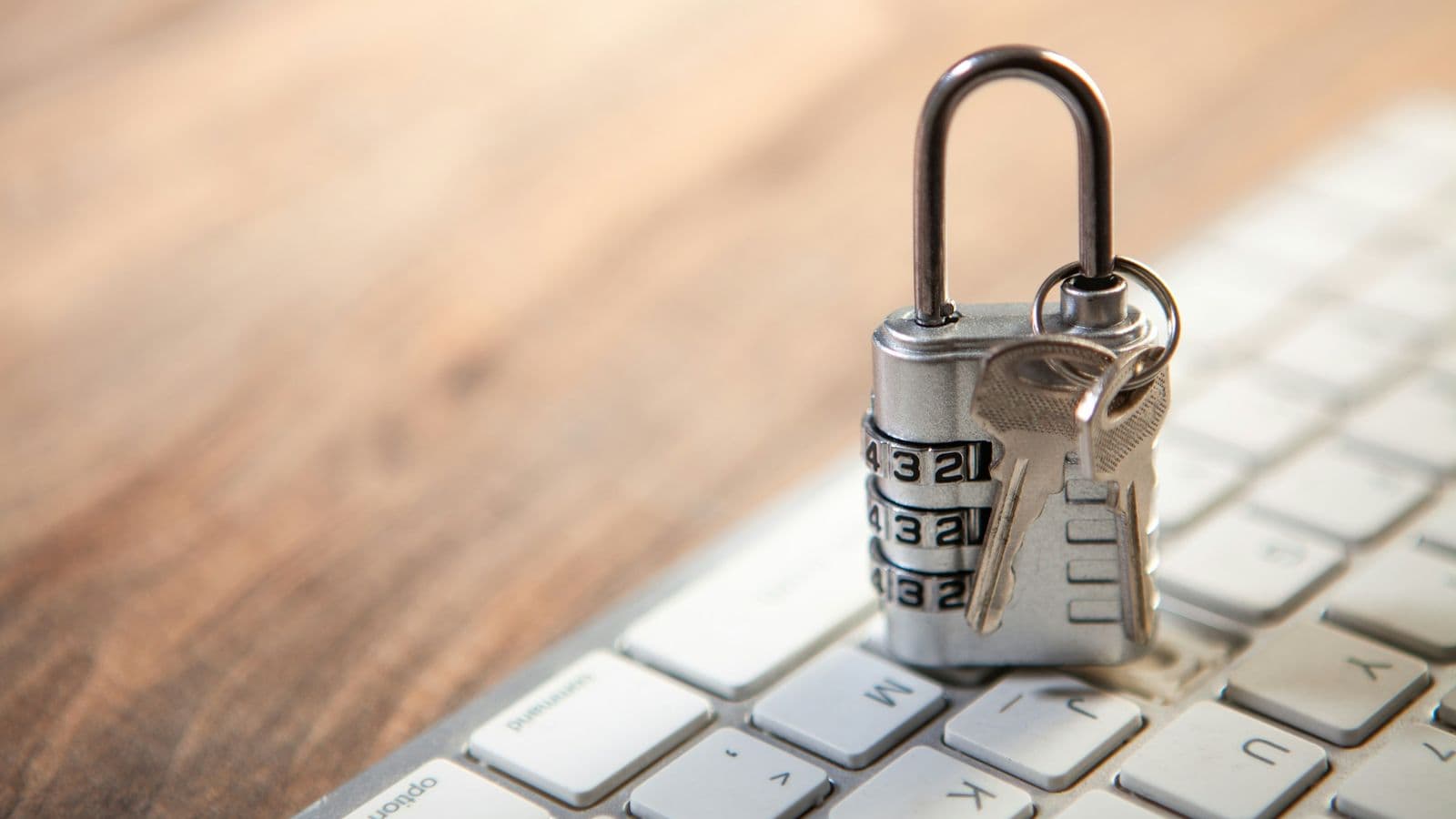 A silver combination padlock with two keys on its ring resting on a white computer keyboard on a wooden desk. Cloud providers secure the infrastructure, but the keys to your own accounts and data are still yours to look after