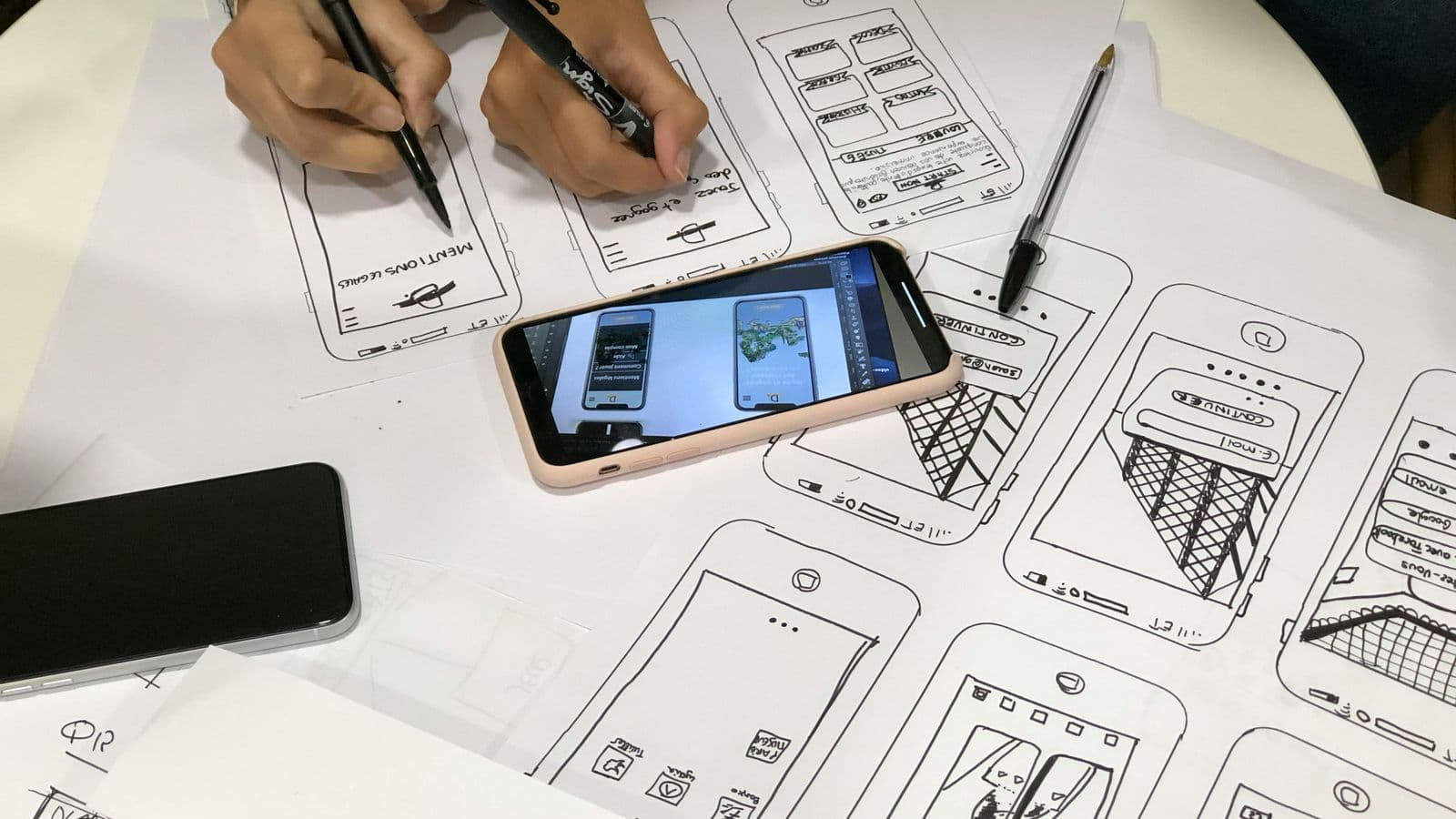 Wireframes on paper beside a phone — interface design runs in parallel with the ledger, never ahead of it