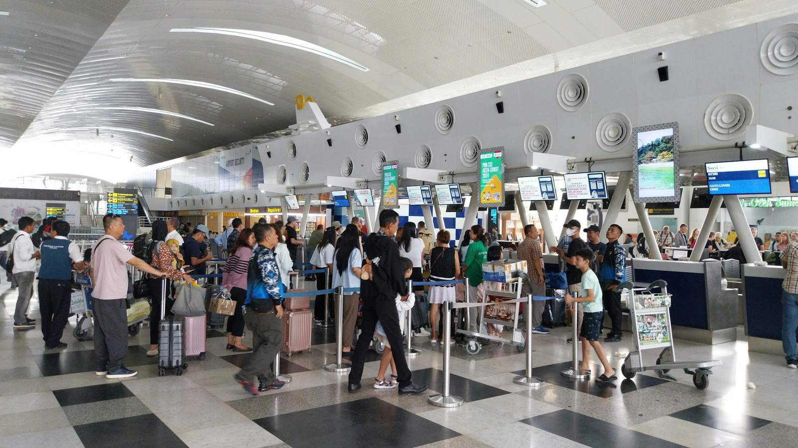 A crowded airport check-in hall with travellers and luggage trolleys queued between barriers, waiting for a long row of counters. A load balancer does the job of the person who sends each traveller to the next free counter