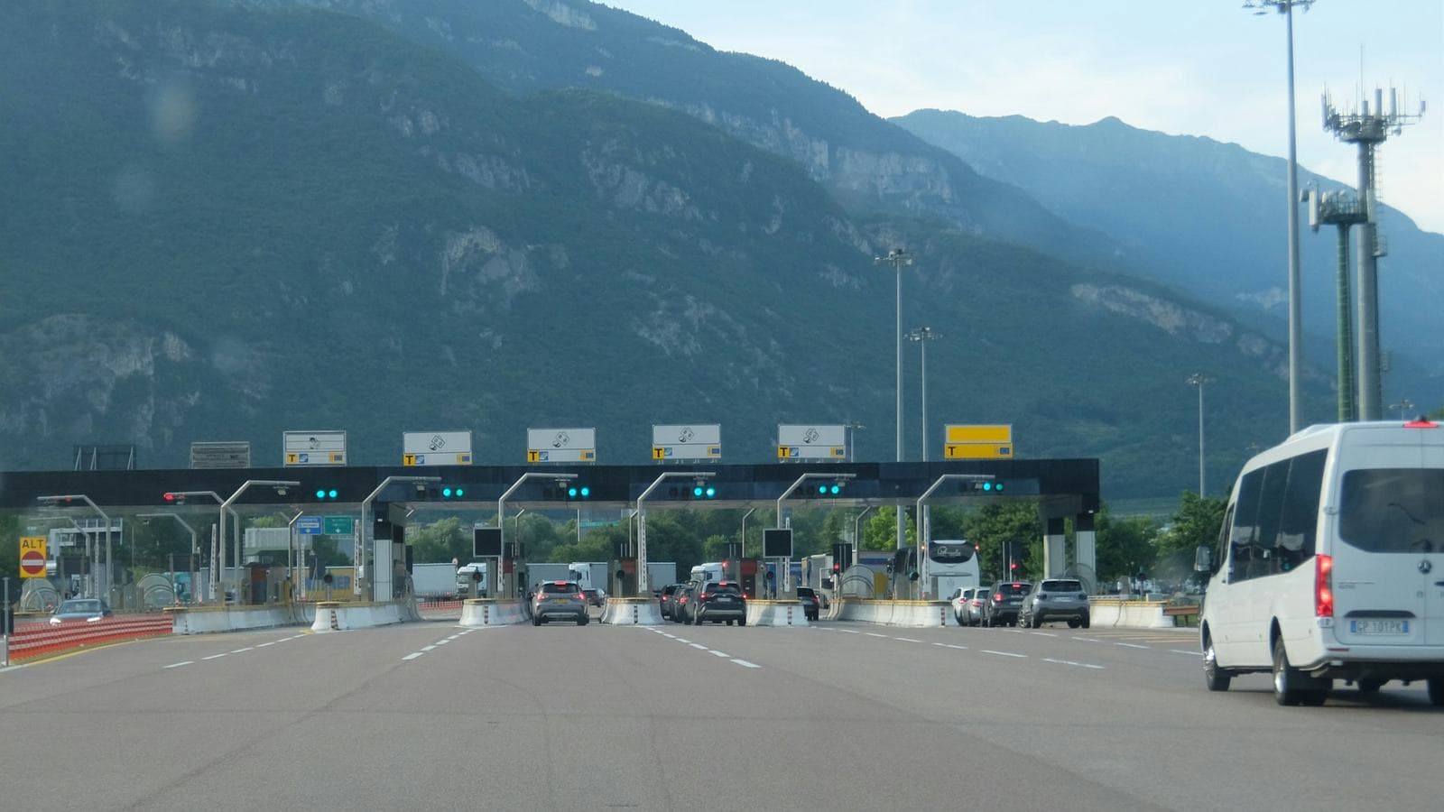 Cars approaching a wide toll plaza with a long row of open booths under a row of green signals, mountains behind. A load balancer works the same way, spreading incoming traffic across many servers so no single lane backs up