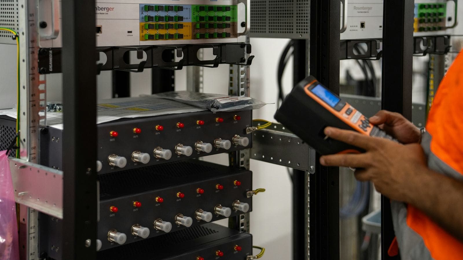 A technician in a high-visibility vest holding a handheld diagnostic meter up to a server rack full of network ports and patch panels. Health checks are a load balancer's automated version of this routine inspection, run every few seconds on every server