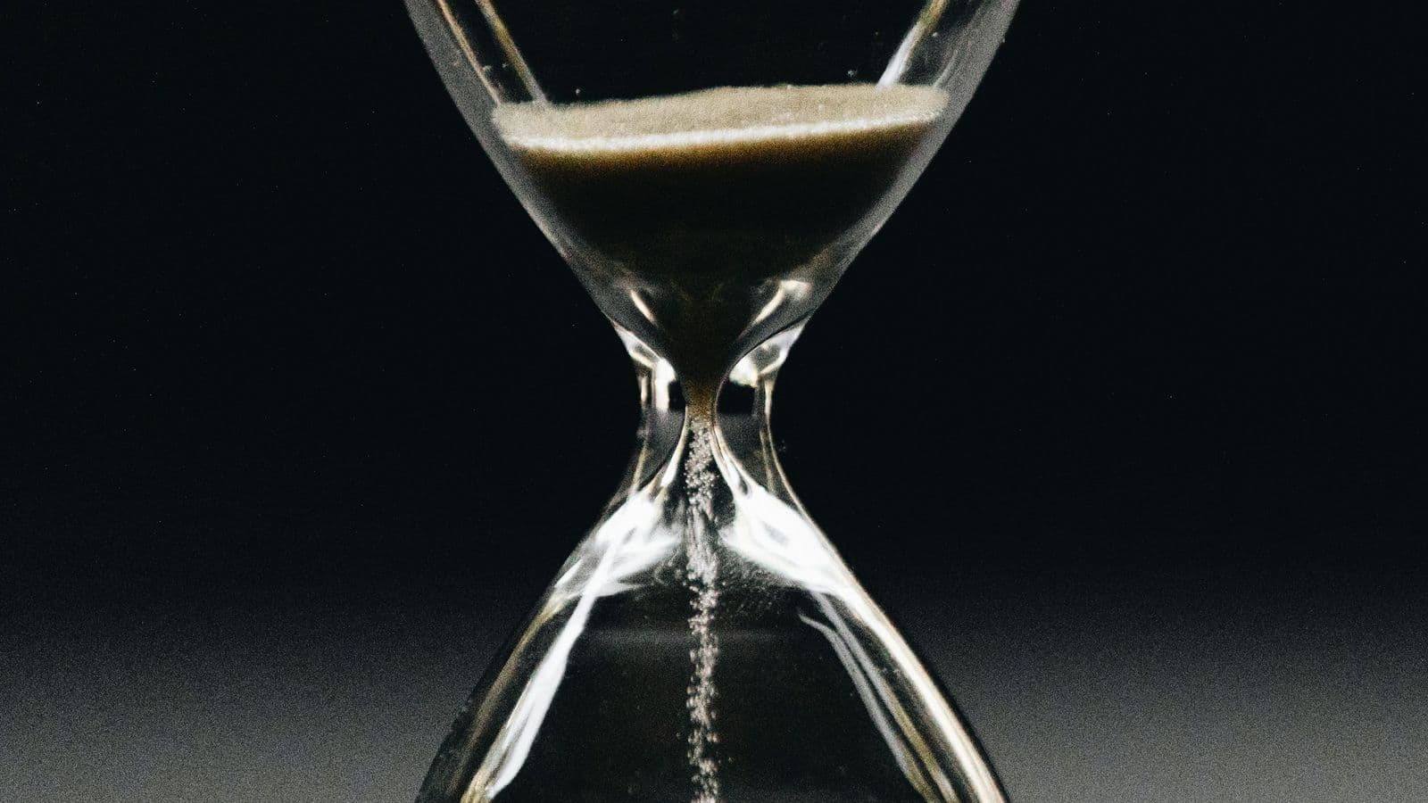A glass hourglass against a black background with sand trickling through the narrow neck, a reminder that every second a page takes to load is time a visitor spends deciding whether to leave