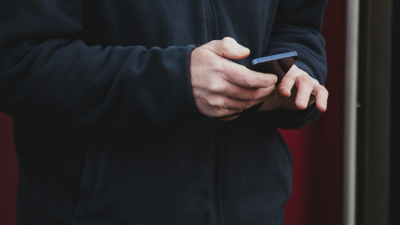 A person in a dark jacket holding a smartphone in both hands, thumb on the screen. Most visitors meet a local business's website on a phone like this, often on mobile data rather than Wi-Fi