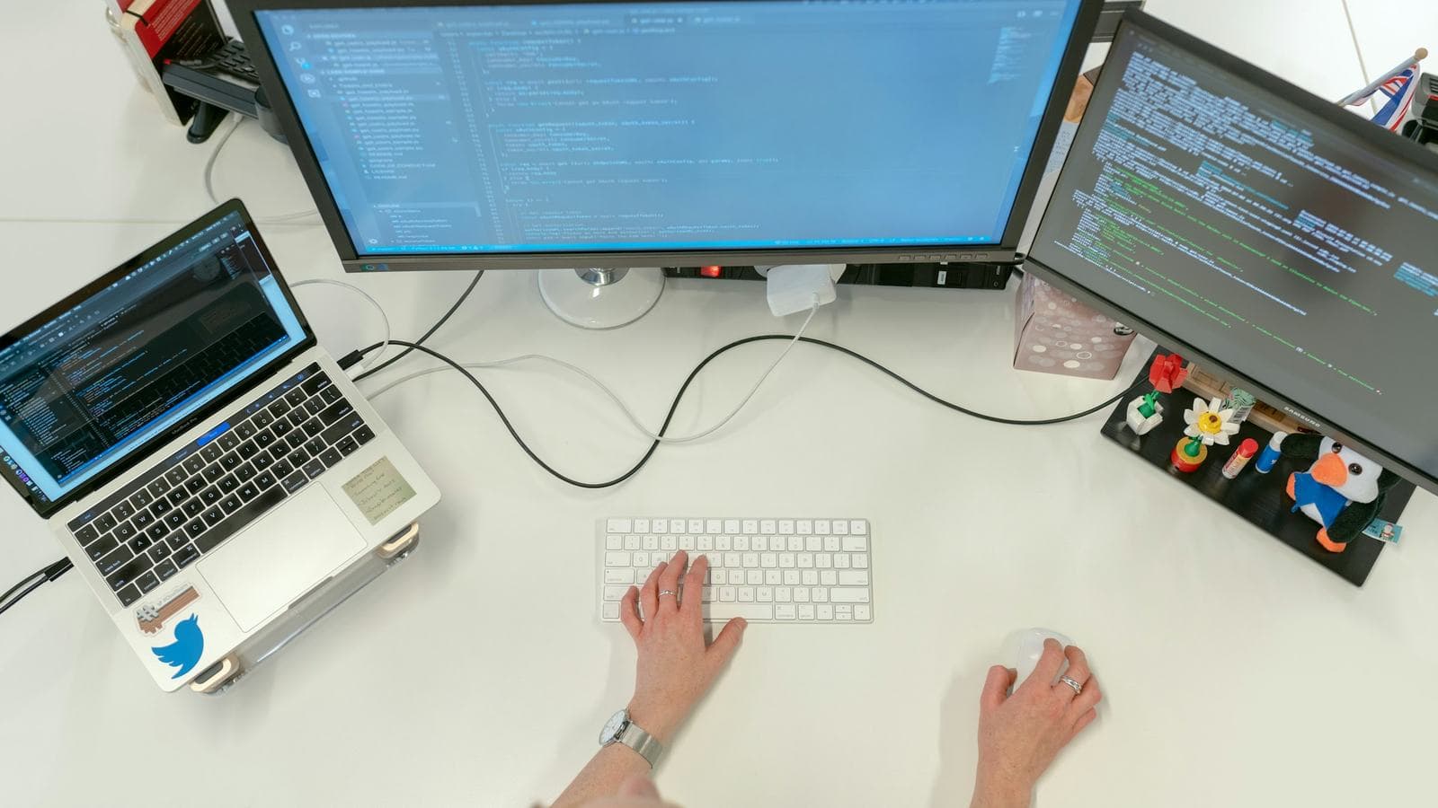A desk seen from above with a laptop, two monitors full of code, a keyboard and a notebook. Getting the project running on your own machine is often the first hurdle of an internship