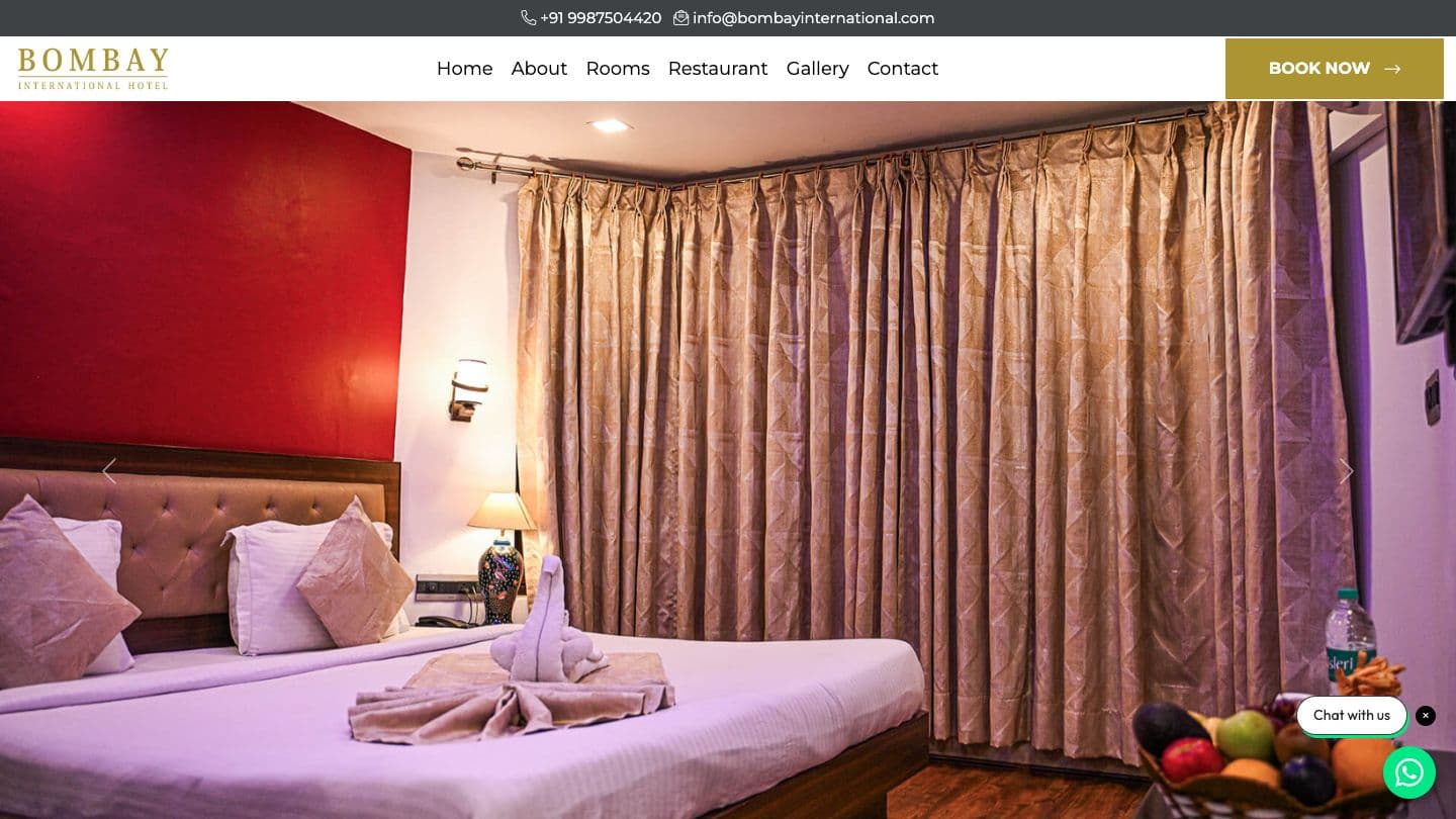 Bombay International Hotel homepage with a gold Book Now button in the header and a full-width photo of a hotel room with a red feature wall, a double bed and a towel swan